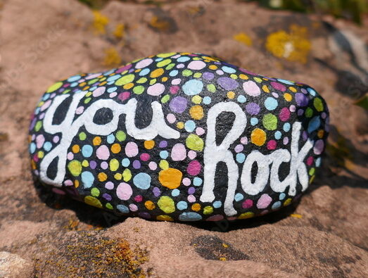 May I have alt text for this A photo of a smooth, dark stone painted with colorful polka dots in white, yellow, blue, pink, purple, and green, with "you Rock" hand-lettered across the center in white. The rock sits on a larger sandstone boulder with green leafy plants blurred in the background.